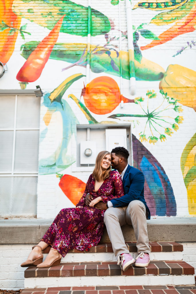 Couple sitting, hugging and laughing near Whiskey Bird in Midtown Atlanta Georgia in front of a mural of vegetables | Photo by Meli and Chris Wedding Photography