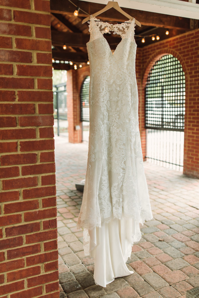 Long lace wedding dress hanging from a hanger next to brick walls at the Cotton Gin at Mill Creek.. Photo by Meli and Chris Wedding Photography.