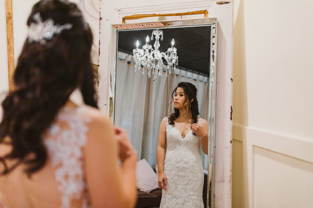Bride looking at herself in the mirror while fixing her hair with a chandelier in the background, inside the Cotton Gin at Mill Creek. Photo by Meli and Chris Wedding Photography.