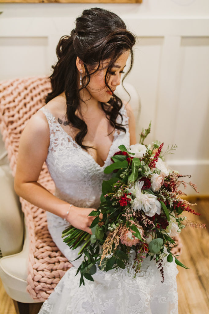 Bride sitting down wearing a sleeveless lace wedding dress looking down at her flower bouquet. inside the Cotton Gin at Mill Creek. Photo by Meli and Chris Wedding Photography atlweddingphotos.com